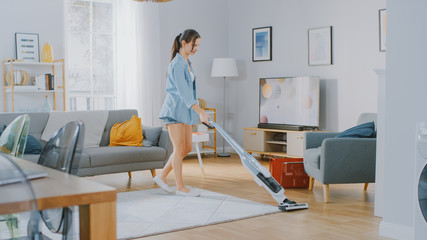 Young Beautiful Woman in Jeans Shirt and Shorts is Vacuum Cleaning a Carpet in a Bright Cozy Room...