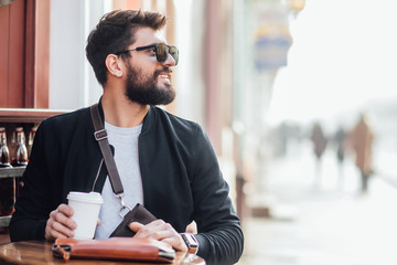 Stylish bearded man during morning walk with cup of coffe near street cafe