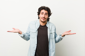 Young handsome man against a white background doubting and shrugging shoulders in questioning...