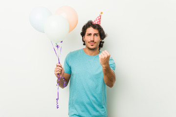 Young man celebrating a party holding balloons showing fist to camera, aggressive facial expression.