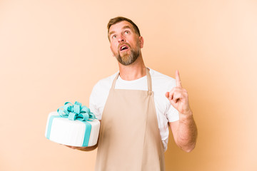 Senior holding a cake isolated on beige background pointing upside with opened mouth.