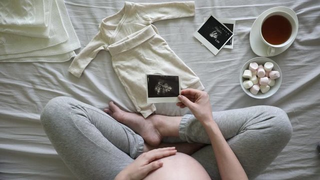 Top vew of expecting pregnant woman touching her belly while looking at ultrasound scans at home, pregnancy and birth concept