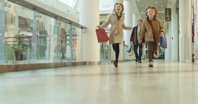 Happy Caucasian kids sister and brother running shopping mall passage with packets and parents following them by walking. Cheerful children having fun in store with mother and father.