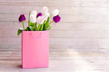 Spring flowers. Blooming tulips in pink shopping bag at wooden background. White, lilac and purple bouquet. Still life in morning sun light. Presents for holidays. Copy space.