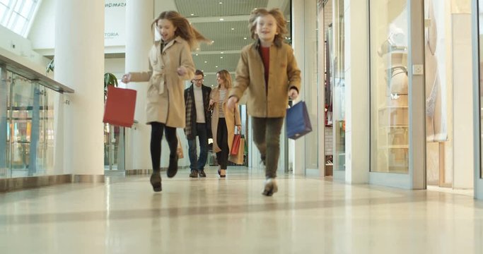 Joyful Caucasian kids sister and brother running shopping mall passage with packets and parents following them by walking. Happy boy and girl having fun in store with mother and father.