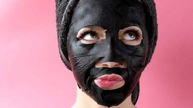 Young woman apply black cosmetic fabric facial mask on pink background. Face peeling mask with charcoal, spa beauty treatment, skincare, cosmetology. Close up