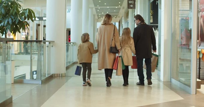 Rear of Caucasian family with kids spending weekend in shopping mall. Smiled parents walking hand in hand with son daughter with bags. Back view on little sister and brother with mother and father.