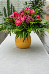 a brown table with a white tablecloth stands a colourful bouquet of flowers in a yellow vase