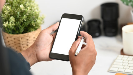 Photographer's hands holding white blank screen mobile and pen at working desk.
