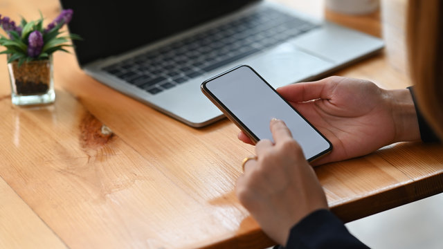 Cropped image of businesswoman's hands holding a white blank screen smartphone at the wooden working desk.