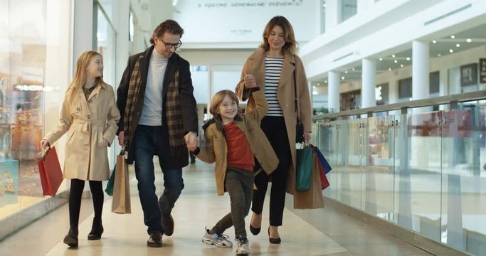 Caucasian family with two kids spending weekend in shopping mall. Smiled parents walking hand in hand with small son and daughter with bags from shop. little sister and brother with mother and father.