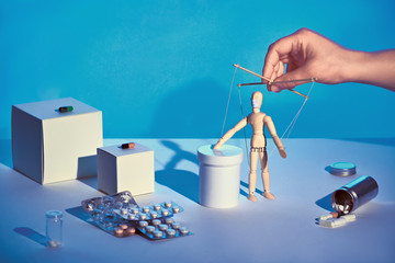 Hooked on medicine. Sinister hand controlling wooden puppet among various medications, pills,...