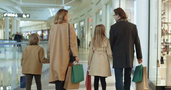 Rear of Caucasian family with kids spending weekend in shopping mall. Smiled parents walking hand in hand with son daughter with bags. Back view on little sister and brother with mother and father.