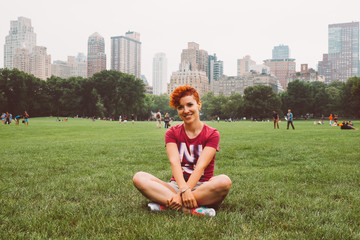 Happy woman sitting on grass, enjoying vacation outdoors in Central Park, New York City, USA. Smiling young tourist girl in summer sun relaxing in Manhattan, visiting NY.