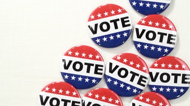 Looking down slider view of round red, white and blue VOTE buttons to I VOTED TODAY stickers and a roll of those stickers on a white linen table cloth