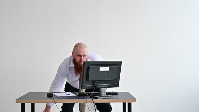 Angry bald man with a red beard in the office in a business suit smashes an ax with a computer. The manager with a nervous breakdown, breaks the monitor.