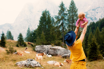 Back of a mother outside in nature lifting up her happy baby in front of a mountain meadow with cows