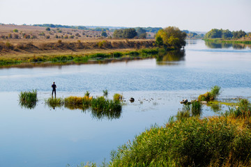 Man traveler silhouette near river with green forest hills. Travel outdoor lifestyle. Fisherman standing in grass, fishing and enjoy autumn nature. Rural landscape with blue lake and man catch fish.