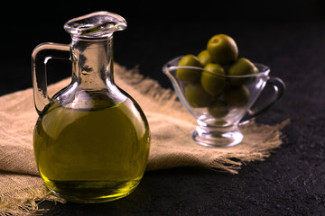  Olive oil in a jug on a black background and olives in a sauceboat in the background. The concept...
