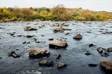 Clear blue river, lush green rural forest and rocky stone cascade in warm sunlight. Colorful autumn landscape, Southern Bug, Ukraine. Beautiful fall nature with green trees. Mountain hills view.