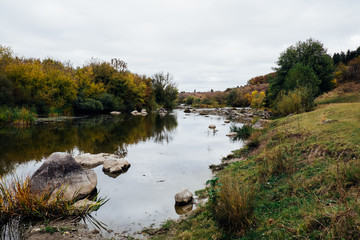 Meandering river, lush green rural forest and rocky stone cascade in warm sunlight. Colorful autumn landscape, Southern Bug, Ukraine. Beautiful fall nature with yellow trees. Mountain hills view.