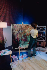 A young female artist is creating an abstract painting. She is working in the loft space with dimmed lights. She is using oil paint and a brush.