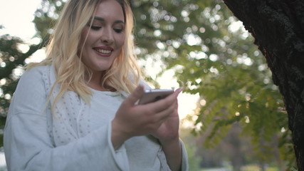 Naklejka premium Pretty Woman Plus Size Hold Mobile Park Background. Blonde Long Haired Girl Browse Smartphone Use Digital Technology Outdoors. Attractive Caucasian Lady Look Around Stand by Tree