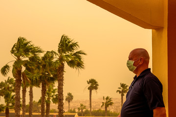 A man is standing on a balcony in the hotel and has a face mask to protect him from the Calima sandstorm, which brings a lot of fine dust. Concept: health and travel