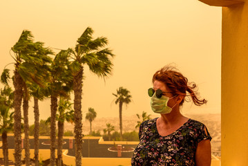 A woman stands on a balcony in the hotel and has a face mask and sunglasses to protect her from the Calima sandstorm, which brings a lot of fine dust. Concept: health and travel