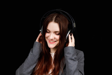 Fototapeta premium Young pretty woman listening to music in headphones. Girl in grey jacket on black background. Beautiful girl with big earphones