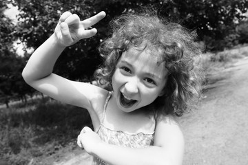 Happy, funny, crazy little girl having fun outdoors in the summer. Adorable, cute curly child, kid fooling, making funny faces, grimaces on beautiful summer day. Black and white photo.