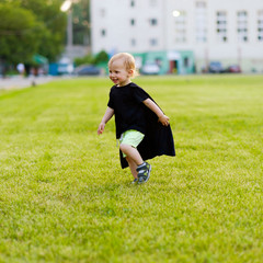 Happy boy running on green meadow. Little kid in black hero cloak running and smiling in the field on summer day outside in the park. Happy childhood concept. Cute child plays superhero outdoor.