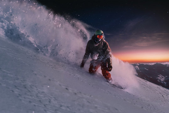 night skating snowboarder curved and brakes spraying loose deep snow on freeride slope under the starry sky and sunset light