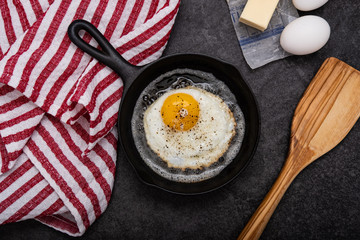 Single Fried Egg, Sunny Side Up in cast iron skillet with a wooden spatula, kitchen towel, on a slate countertop