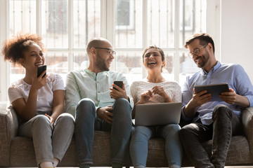 Obraz premium Cheerful multiracial friends using diverse gadgets sitting together on couch