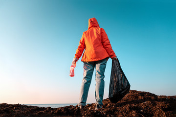 A young Caucasian female volunteer in a jacket poses with a large garbage bag and a plastic bottle in her hands. Rear and bottom view. In the background, the sky and coast line. Copy space