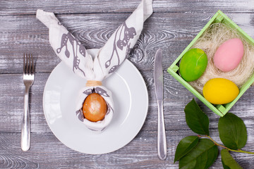 Composition with an Easter egg wrapped in a napkin in the form of rabbit ears on a white plate, cutlery, colorful Easter eggs in a green wooden box on a wooden gray table. Flat lay
