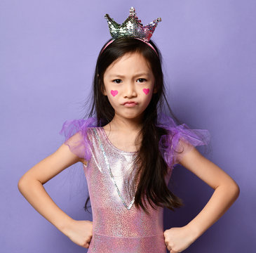 Unhappy Mad Asian Kid Girl In Party Dress, Crown And With A Painted Red Hearts Sign Is Posing With Her Hands On Her Hips