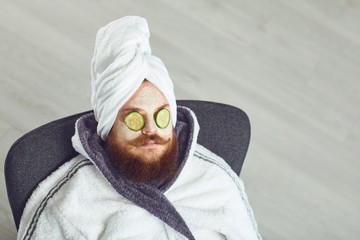 Funny fat bearded man with a cosmetic mask on his face in bathrobe towel on his head on his face resting against a gray background