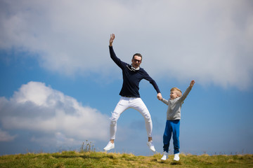 Carefree father and son holding hands while jumping on the field against the sky.