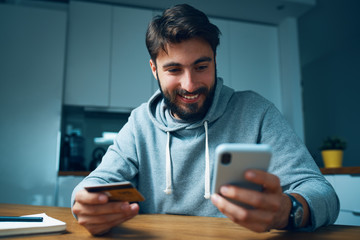 Young man sitting at home late and ordering online with smartphone and credit card