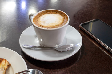A cup of cappuccino, a latte in a cafe on a table with a dark wooden surface, next to a smartphone.