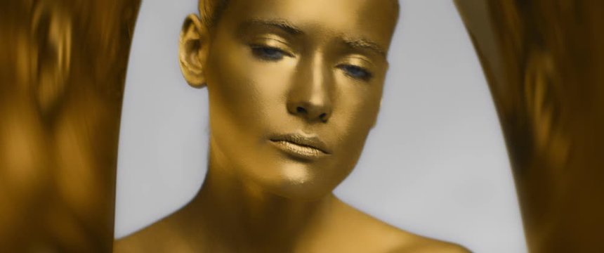 female model with gold trendy makeup and body art open her eyes and looks at camera, fashion trend, blue eyes, bronze or golden skin color or tone, reflective surface, anamorphic shot