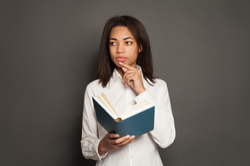 Thinking mixed race ethnicity black woman student in white shirt with book on gray background