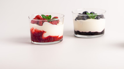 Two layered yogurt desserts. On with raspberry jam and fresh raspberries, another with blueberry jam, fresh blueberries and mint. White background
