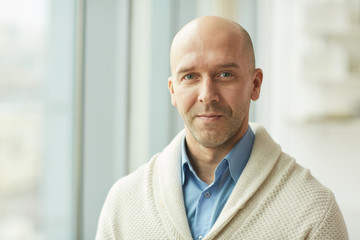 Head and shoulders portrait of balding mature man wearing cardigan looking at camera while standing by window in white office, copy space - obrazy, fototapety, plakaty