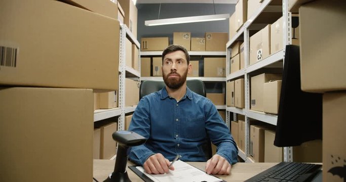 Caucasian man sitting at table with many carton boxes as having a lot of work and parcels in delivery store. Postman tired and busy with cargo and mail for registration and delivering.