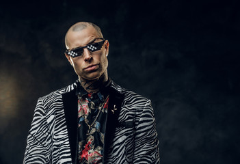 Charming, masculine, tattooed, bald male model posing in a dark studio for the photoshoot wearing fashionable custom made zebra striped style tuxedo, golden chain, rose patterned shirt and black pixel