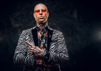 Sreious, stylish, tattooed, bald male model posing in a dark studio for the photoshoot wearing fashionable custom made zebra striped style tuxedo, golden chain, rose patterned shirt and red glasses