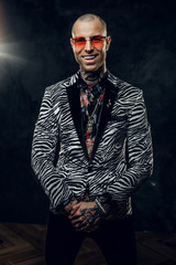 Obraz premium Uroczy, stylowy, wytatuowany, łysy model pozujący w ciemnym studiu do sesji zdjęciowej w modnym, robionym na zamówienie smokingu w paski zebry, złotym łańcuszku, koszuli we wzory w róże i czerwonych okularach